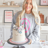 A smiling woman in a gray sweatshirt with a pink circle logo stands in a bright kitchen behind a unicorn cake. Baking supplies are on the counter. Jenna Rae Cakes offers Online Classes to help you create magical cakes like this at home.
