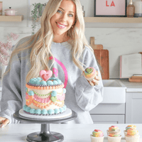 A smiling woman with long blonde hair stands in a kitchen holding a cupcake, with a Jenna Rae Cakes decorated cake and matching cupcakes, highlighting their Online Classes.