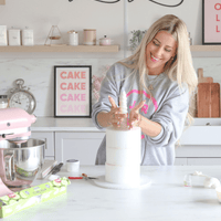 A woman smiles while decorating a tall white cake in a modern kitchen. Baking tools, a pink mixer, and cake supplies are on the counter, with Jenna Rae Cakes Online Classes visible on the wall behind her.