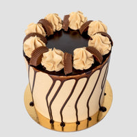 The Peanut Butter Cup Cake by Jenna Rae Cakes is a tall cake with light brown frosting, chocolate drip, swirls of frosting, and halved chocolate peanut butter cups on a gold cake board.
