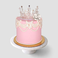 The Princess Crown Cake by Jenna Rae Cakes is a pink birthday cake with white icing bows, pearl decorations, a clear crown topper, and a plaque reading Happy 3rd Birthday, Charlie, displayed on a marble cake stand.