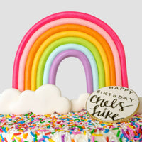 The Rainbow Cake by Jenna Rae Cakes is a colorful cake with rainbow and cloud decorations, rainbow sprinkles, and a round cookie on top that reads Happy Birthday Chels + Luke.