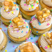 Regular Cupcakes by Jenna Rae Cakes are arranged closely together in gold wrappers and topped with swirls of frosting, colorful confetti sprinkles, and pieces of caramel peanut nougat candy on a white surface.
