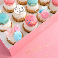A pink box of Jenna Rae Cakes Reveal Works Cupcakes, decorated with pink and blue macarons (one reads He or She?), pastel meringues, and white sprinkles.
