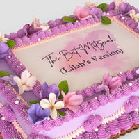 The Sugar Flower Sheet Cake by Jenna Rae Cakes is a rectangular cake with purple and pink frosting, sugar flowers, green leaves, and The Bat Mitzvah (Lilah’s Version) written in elegant black script on a white center panel.