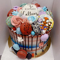 The Kids Works Cake by Jenna Rae Cakes is a colorful birthday cake topped with meringues, macarons, candies, chocolate drip, and a Spider-Man cookie. “Happy 3rd Birthday Callum” is written in black icing on top.