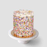 The Vanilla Sprinkle Cake by Jenna Rae Cakes is a round cake covered in colorful rainbow sprinkles with white frosting swirls, displayed on a white marble cake stand against a plain light gray background.