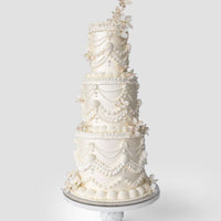 The Wedding Cakes $1,000+ by Jenna Rae Cakes features four tiers of white cake with piped icing, scalloped designs, and delicate sugar flowers, showcased on a marble stand against a simple light backdrop.