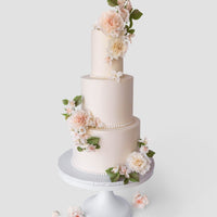 A three-tiered white Wedding Cake by Jenna Rae Cakes ($1,000+) features lifelike pale pink and white flowers with green leaves, presented on a white cake stand with additional flowers arranged at the base.