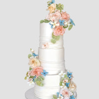 The Wedding Cakes $1,000+ by Jenna Rae Cakes features a three-tiered white cake adorned with pastel pink, peach, blue, and white flowers plus green leaves, displayed on a white stand against a simple backdrop.