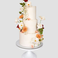 A three-tiered white Wedding Cakes $1,000+ by Jenna Rae Cakes, adorned with realistic red, orange, and white sugar flowers, is displayed on a white cake stand against a plain background.