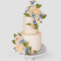 Jenna Rae Cakes Wedding Cakes Under $1,000 features a two-tiered white cake adorned with pastel pink, blue, and white flowers plus green leaves, displayed on a marble stand against a plain light background.