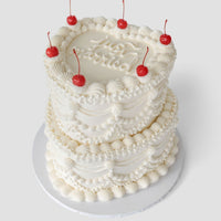 A two-tiered white cake decorated with ornate piped frosting, topped with six red cherries and the words Not Patriotic written on the top layer, set on a round white cake board against a plain background.