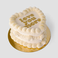 A heart-shaped cake from Jenna Rae Cakes’ Wedding Cakes Under $200 features white frosting, piped edges, and love you more in gold on top, set on a gold board against a simple backdrop.