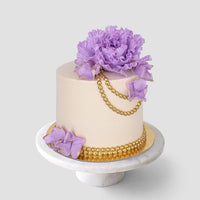 A beige-frosted cake from Jenna Rae Cakes’ Wedding Cakes Under $200, set on a white stand and adorned with gold beads and large purple sugar flowers on top and the side.