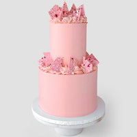 The Wedding Cakes Under $500 by Jenna Rae Cakes features a two-tiered pink cake with pink frosting swirls, pink chocolate shards, and sprinkles, shown on a white stand against a simple backdrop.