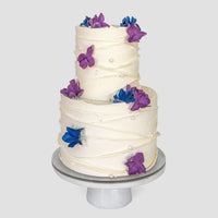 From Jenna Rae Cakes Wedding Cakes Under $500 collection: a two-tiered cake with white frosting, decorated with purple and blue flowers and pearl-like accents, displayed on a round white stand against a light gray background.