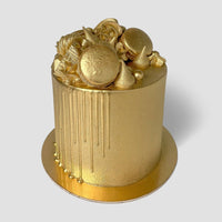 A round cake glistens with Jenna Rae Cakes’ Antique Gold Edible Lustre Dust, featuring metallic gold frosting, gold drips, macarons, candies, and decorations on a matching gold board set against a plain gray backdrop.