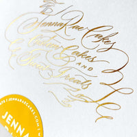 A white card with elegant gold cursive lettering reads Jenna Rae Cakes. In the bottom left, a partial yellow sticker shows the name Jenna Rae. Displayed with the Cookie Sandwich Gift Box from Jenna Rae Cakes.