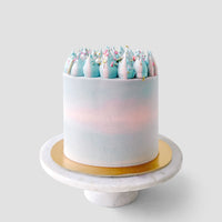 The Cotton Candy Cake by Jenna Rae Cakes is a tall, round cake with pastel blue and pink frosting, swirled dollops, and colorful sprinkles. It sits on a gold cake board and white marble stand against a plain gray background.