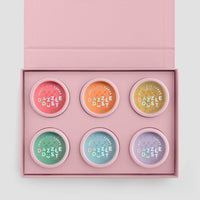 The Dazzle Dust Box by Jenna Rae Cakes is a pink box with “MAKE LIFE DAZZLE” on the lid, featuring six pastel-colored circular tins labeled “DAZZLE DUST,” arranged in two neat rows inside.