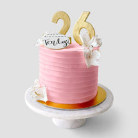 The Design Your Own Cake by Jenna Rae Cakes features pink frosting, gold 26 toppers, white flower decorations, and a Happy Birthday Tenley card, displayed on a marble cake stand.