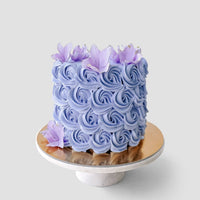 The Design Your Own Cake by Jenna Rae Cakes features a round design with intricate lavender buttercream rosettes and purple flowers, displayed on a marble cake stand against a plain light gray background.