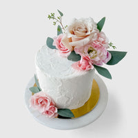 The Fresh Floral Buttercream Cake by Jenna Rae Cakes features pink and cream flowers with green leaves on white frosting, presented on a round white marble stand with a gold base against a light background.