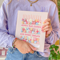 A woman in a lavender sweater and jeans holds the Jenna Rae Cakes and Sweet Treats Cookbook by Jenna Rae Cakes, featuring a pastel cover with macarons and candies. She has manicured nails and layered gold jewelry.