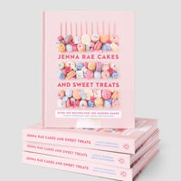 A stack of four Jenna Rae Cakes and Sweet Treats Cookbooks by Jenna Rae Cakes, with one standing upright on top, displays colorful macarons and desserts on the pink covers against a light grey background.