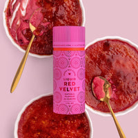 Three ramekins of red velvet crème brûlée with caramelized tops and gold spoons are arranged around a pink bottle of Jenna Rae Cakes Liquid Red Velvet natural flavoring on a pink background.