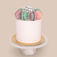 The Macaron Crown Cake by Jenna Rae Cakes features white frosting, colorful macarons, and a Happy Birthday sign, displayed on a round marble cake stand against a beige background.