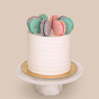 The Macaron Crown Cake by Jenna Rae Cakes features a white frosted finish topped with colorful macarons, displayed on a gold cake board and white marble stand, set against a plain beige background.