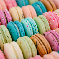The Macaron Gift Box by Jenna Rae Cakes features rows of colorful pastel macarons in pink, blue, green, and beige, some topped with decorative sprinkles and beautifully arranged for a delightful presentation.