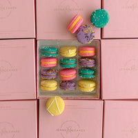 A Jenna Rae Cakes Macaron Gift Box featuring yellow, purple, green, and pink macarons sits atop branded pink boxes, with three macarons displayed outside the container.
