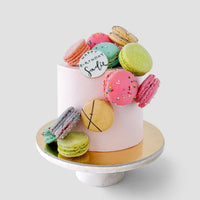 The Macaron Lovers Cake by Jenna Rae Cakes is a white frosted birthday cake adorned with colorful macarons and a white fondant plaque reading Happy Birthday Sadie, presented on a white stand with a gold base.