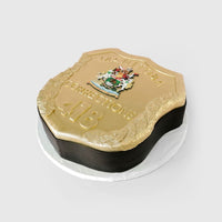 A cake shaped like a gold Manitoba Corrections badge, featuring the Manitoba coat of arms, the numbers “418,” and decorative gold detailing, displayed on a white round base.