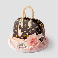 A cake designed to look like a Louis Vuitton handbag, decorated with gold monogram patterns, beige handles, a pink fondant flower, and a “Happy Birthday Mifsara” tag on a pale pink base.