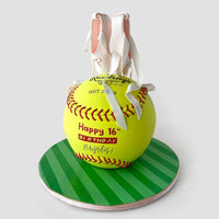 A round cake decorated as a yellow softball with red stitching sits on a green striped base. White fondant ballet slippers with pink accents rest on top. The cake reads Happy 16th Birthday Bristol! in red and yellow.
