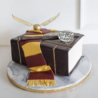 A cake designed to look like a book, decorated with a maroon and gold scarf, a golden snitch, and a plaque that reads Happy Birthday Jessica! on a round marble-patterned board.