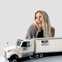 A smiling woman with long blonde hair sits behind a detailed cake shaped like a white Meyer Bros. semi truck and trailer. The background is plain gray.