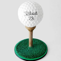 A realistic cake shaped like a golf ball on a wooden tee, set on a circular base decorated to look like green grass, with Titleist 28 written on the golf ball.