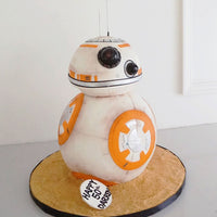 A cake shaped like BB-8, the droid from Star Wars, with white and orange details. It sits on a round sand-colored base with a small sign reading Happy 5th Birthday, Darcey.