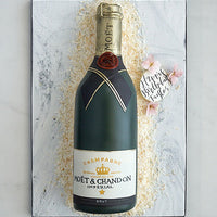 A cake decorated to look like a Moët & Chandon champagne bottle, lying on a white surface with shredded white decorations and a small note reading Happy Birthday Taylor beside pink flowers.