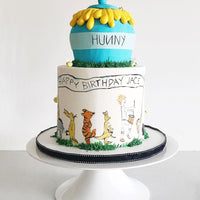 Two-tier birthday cake featuring a blue honey pot on top and drawings of Winnie the Pooh characters around the lower tier, with Happy Birthday Jace written on the side. The cake sits on a white stand.