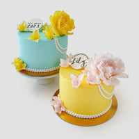 Two Jenna Rae Cakes Peony and Pearls Cakes: one blue with yellow flowers, pearls, and a 2021 topper; the other yellow with pink flowers, pearls, and a Congratulations Lily sign—both on gold cake boards against a white background.