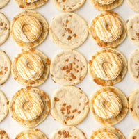 Jenna Rae Cakes Regular Cookie Sandwiches are displayed on parchment paper, featuring plain cookies, some with caramel bits, and others topped with swirled white and caramel-colored frosting.