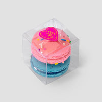 Jenna Rae Cakes’ Signature 2-Macaron Box features a clear plastic container with pink and blue macarons stacked inside, topped with colorful sprinkles and a pink heart sticker, set against a light gray background.