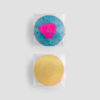 Two macarons from the Jenna Rae Cakes Signature 2-Macaron Box sit on a light gray background—one blue with a pink heart sticker, the other yellow with an embossed circular logo on its wrapper.