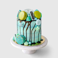 The Full Works Cake by Jenna Rae Cakes is a tall, blue-green ombre cake with chocolate drip, topped with assorted macarons, meringues, and cookies, displayed on a white cake stand against a plain background.
